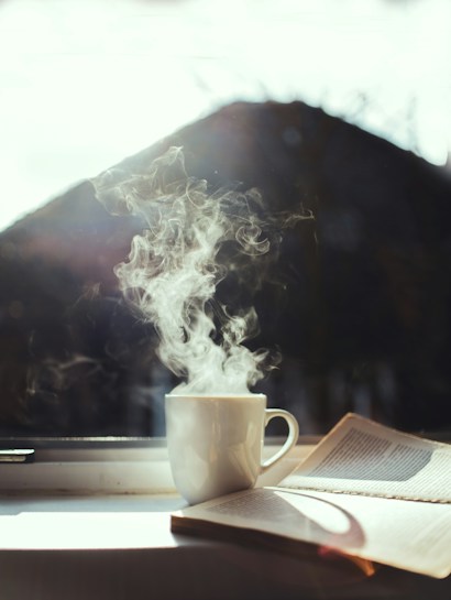 A steaming cup of coffee sitting on a windowsill beside an open book. Sunlight shines through the window, casting soft shadows.