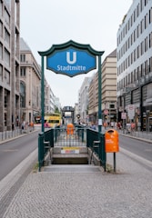 An urban street scene featuring the entrance to a subway station named 'Stadtmitte' with a prominent blue sign displaying a 'U' for underground. People are walking on the sidewalks, and buses can be seen in the background. The street is lined with modern buildings, and the area appears to be a bustling city center.