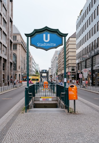 An urban street scene featuring the entrance to a subway station named 'Stadtmitte' with a prominent blue sign displaying a 'U' for underground. People are walking on the sidewalks, and buses can be seen in the background. The street is lined with modern buildings, and the area appears to be a bustling city center.