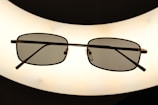 A pair of rectangular framed eyeglasses with dark lenses is centered against a bright, oval-shaped light source, creating a silhouette effect. The frame is thin and metallic, giving a sleek appearance.
