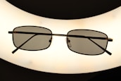 A pair of rectangular framed eyeglasses with dark lenses is centered against a bright, oval-shaped light source, creating a silhouette effect. The frame is thin and metallic, giving a sleek appearance.