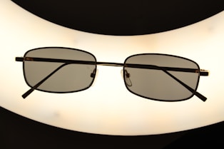 A pair of rectangular framed eyeglasses with dark lenses is centered against a bright, oval-shaped light source, creating a silhouette effect. The frame is thin and metallic, giving a sleek appearance.