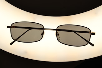 A pair of rectangular framed eyeglasses with dark lenses is centered against a bright, oval-shaped light source, creating a silhouette effect. The frame is thin and metallic, giving a sleek appearance.