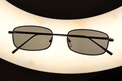 A pair of rectangular framed eyeglasses with dark lenses is centered against a bright, oval-shaped light source, creating a silhouette effect. The frame is thin and metallic, giving a sleek appearance.