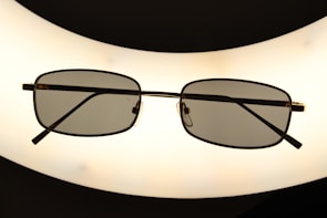 A pair of rectangular framed eyeglasses with dark lenses is centered against a bright, oval-shaped light source, creating a silhouette effect. The frame is thin and metallic, giving a sleek appearance.