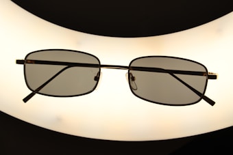 A pair of rectangular framed eyeglasses with dark lenses is centered against a bright, oval-shaped light source, creating a silhouette effect. The frame is thin and metallic, giving a sleek appearance.