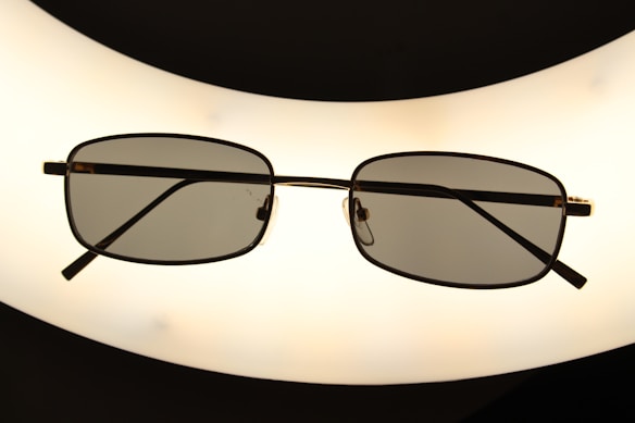 A pair of rectangular framed eyeglasses with dark lenses is centered against a bright, oval-shaped light source, creating a silhouette effect. The frame is thin and metallic, giving a sleek appearance.