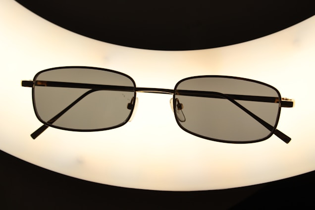 A pair of rectangular framed eyeglasses with dark lenses is centered against a bright, oval-shaped light source, creating a silhouette effect. The frame is thin and metallic, giving a sleek appearance.