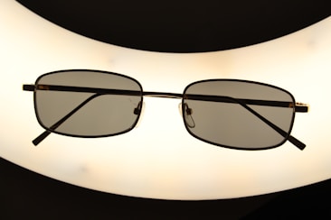 A pair of rectangular framed eyeglasses with dark lenses is centered against a bright, oval-shaped light source, creating a silhouette effect. The frame is thin and metallic, giving a sleek appearance.