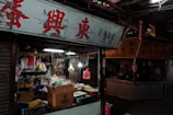 A street market stall is filled with various products including packaged goods, boxes, and hanging bags of snacks. The area appears dimly lit, with fluorescent lights inside the stall and a sign with red Chinese characters above.