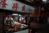 A street market stall is filled with various products including packaged goods, boxes, and hanging bags of snacks. The area appears dimly lit, with fluorescent lights inside the stall and a sign with red Chinese characters above.