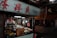 A street market stall is filled with various products including packaged goods, boxes, and hanging bags of snacks. The area appears dimly lit, with fluorescent lights inside the stall and a sign with red Chinese characters above.