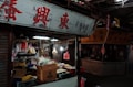A street market stall is filled with various products including packaged goods, boxes, and hanging bags of snacks. The area appears dimly lit, with fluorescent lights inside the stall and a sign with red Chinese characters above.