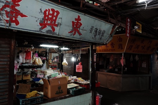 A street market stall is filled with various products including packaged goods, boxes, and hanging bags of snacks. The area appears dimly lit, with fluorescent lights inside the stall and a sign with red Chinese characters above.