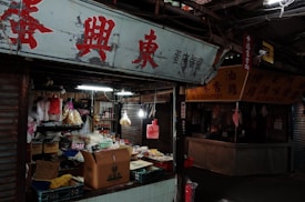 A street market stall is filled with various products including packaged goods, boxes, and hanging bags of snacks. The area appears dimly lit, with fluorescent lights inside the stall and a sign with red Chinese characters above.