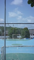 A small, enclosed soccer field with a blue playing surface is surrounded by a tall chain-link fence. The goalpost is visible at the far end, and there are trees and buildings in the background under a partly cloudy sky.