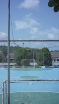 A small, enclosed soccer field with a blue playing surface is surrounded by a tall chain-link fence. The goalpost is visible at the far end, and there are trees and buildings in the background under a partly cloudy sky.