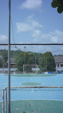 A small, enclosed soccer field with a blue playing surface is surrounded by a tall chain-link fence. The goalpost is visible at the far end, and there are trees and buildings in the background under a partly cloudy sky.