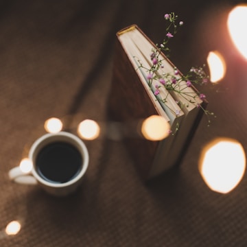 A hardcover book is positioned upright with small purple flowers peeking between its pages. Nearby, there's a white cup filled with dark coffee. Warm, blurred lights are scattered around, creating a cozy ambiance.
