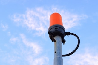 A tall metal pole with a bright orange beacon light mounted at the top, set against a clear blue sky with wispy clouds. A black cable is attached to the beacon and runs down the pole.