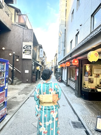 A person wearing a colorful kimono walks down a narrow street lined with traditional buildings and red lanterns. The scene appears peaceful and culturally vibrant, capturing the essence of a historic Japanese setting.