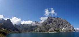 Panoramic view of a serene mountain landscape with rugged peaks and lush green valleys. A calm blue lake lies in the foreground, reflecting the clear blue sky and occasional fluffy clouds. The scene is dominated by large rocky mountains, creating a sense of grandeur and tranquility.