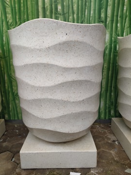 A large, textured beige planter with a wavy pattern is placed on a square platform. The background features a bamboo-themed backdrop, contributing to a natural and serene setting. The planter appears to be made of a stone-like material.