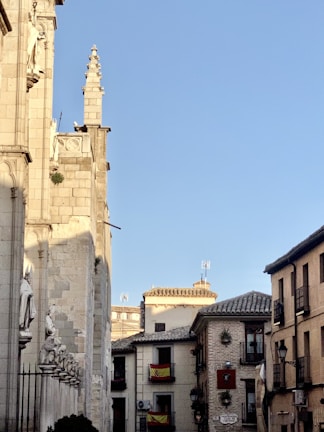 A narrow street lined with traditional European buildings, one of which is an ornate stone structure adorned with statues. Spanish flags hang from balconies, and the architecture features historical elements like spires and clay roof tiles. The scene is set under a clear blue sky, lending a tranquil atmosphere to the historic setting.