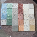 A collection of terrazzo tiles arranged in a grid, showcasing a variety of colors and patterns. The tiles feature multicolored chips embedded in different shades, including green, blue, pink, red, and beige. The background surface is also a speckled terrazzo texture.
