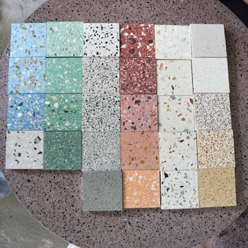 A collection of terrazzo tiles arranged in a grid, showcasing a variety of colors and patterns. The tiles feature multicolored chips embedded in different shades, including green, blue, pink, red, and beige. The background surface is also a speckled terrazzo texture.