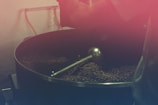 A large coffee roasting drum filled with coffee beans being processed. The setup includes a metal agitator inside the drum, and the beans have a dark brown color. The image has a warm reddish tint, giving it a rustic and vintage feel.
