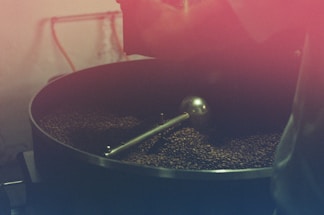 A large coffee roasting drum filled with coffee beans being processed. The setup includes a metal agitator inside the drum, and the beans have a dark brown color. The image has a warm reddish tint, giving it a rustic and vintage feel.