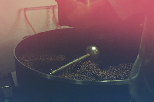 A large coffee roasting drum filled with coffee beans being processed. The setup includes a metal agitator inside the drum, and the beans have a dark brown color. The image has a warm reddish tint, giving it a rustic and vintage feel.