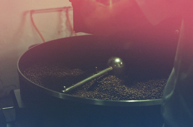 A large coffee roasting drum filled with coffee beans being processed. The setup includes a metal agitator inside the drum, and the beans have a dark brown color. The image has a warm reddish tint, giving it a rustic and vintage feel.