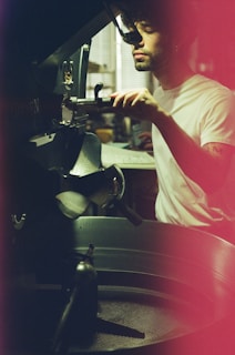 A person operating a machine in a dimly lit setting, wearing a white t-shirt with focused attention. The scene includes industrial equipment, possibly involving coffee roasting, and has a warm, vintage filter effect.