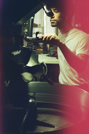 A person operating a machine in a dimly lit setting, wearing a white t-shirt with focused attention. The scene includes industrial equipment, possibly involving coffee roasting, and has a warm, vintage filter effect.