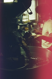 A person appears to be operating a coffee roasting machine, with roasted coffee beans spilling out into a metal container. The setting is dimly lit, with a warm ambiance and a slightly blurred background that suggests a café or industrial space. The person is focused and engaged in the coffee roasting process.