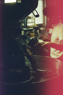A person appears to be operating a coffee roasting machine, with roasted coffee beans spilling out into a metal container. The setting is dimly lit, with a warm ambiance and a slightly blurred background that suggests a café or industrial space. The person is focused and engaged in the coffee roasting process.