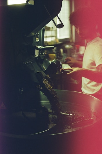 A person appears to be operating a coffee roasting machine, with roasted coffee beans spilling out into a metal container. The setting is dimly lit, with a warm ambiance and a slightly blurred background that suggests a café or industrial space. The person is focused and engaged in the coffee roasting process.
