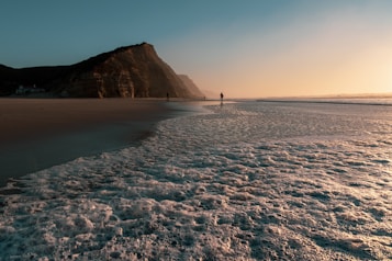 The image features a serene beach at sunset with foamy waves gently reaching the shore. In the background, a large cliff rises against the sky, adding contrast to the smooth horizon. A few silhouettes of people are visible in the distance, enjoying the tranquil setting.