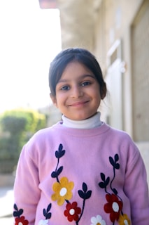 A young girl with dark hair is smiling at the camera. She is wearing a pink sweater with floral patterns in yellow, red, white, and black. The background is slightly blurred and outdoor, possibly depicting a residential area.
