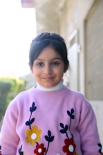 A young girl with dark hair tied back, wearing a light purple sweater adorned with colorful floral designs. She stands in an outdoor setting with a blurred background showing green foliage and a building structure.