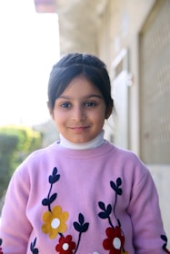 A young girl with dark hair tied back, wearing a light purple sweater adorned with colorful floral designs. She stands in an outdoor setting with a blurred background showing green foliage and a building structure.