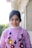 A young girl with dark hair tied back, wearing a light purple sweater adorned with colorful floral designs. She stands in an outdoor setting with a blurred background showing green foliage and a building structure.