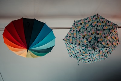 Two umbrellas are hanging upside down from a ceiling. The left umbrella displays vibrant colors arranged in segments, including red, orange, yellow, green, and blue. The right umbrella features a geometric pattern with hexagonal shapes in various shades of pink, blue, yellow, and green.