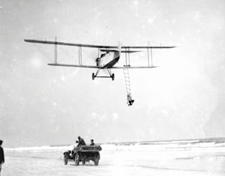 A vintage biplane is flying low above a beach, with a person hanging from a rope ladder attached to the aircraft. Below, a group of people are in an open-top car driving along the sandy shore, possibly in pursuit of or assisting the airborne person.