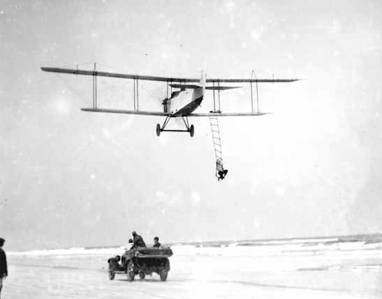 A vintage biplane is flying low above a beach, with a person hanging from a rope ladder attached to the aircraft. Below, a group of people are in an open-top car driving along the sandy shore, possibly in pursuit of or assisting the airborne person.