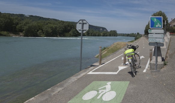 A paved bike path runs beside a calm river, surrounded by lush greenery with a forested hill in the distance. A bicycle with a fluorescent yellow bag parked alongside directional road signs pointing towards nearby locations is seen. Blue sky with scattered clouds is visible above.