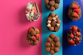 A variety of decorated chocolate eggs are arranged on a split pink and blue background. The eggs are adorned with an assortment of toppings, including strawberries, blackberries, blueberries, assorted chocolates, candy, and marshmallows. Each egg has a unique combination of decorations, with vibrant colors and textures.