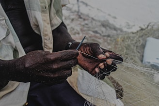 A person is skillfully working with a fishing net, holding a tool in their hands. The focus is on the hands, which are dark and contrasted against the lighter background.